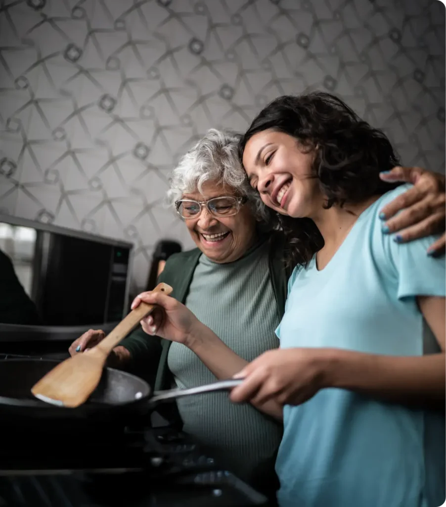 A smiling caregiver and an elderly woman cooking together in a kitchen, illustrating the client's independence and empowered choices within the CDPAP program.